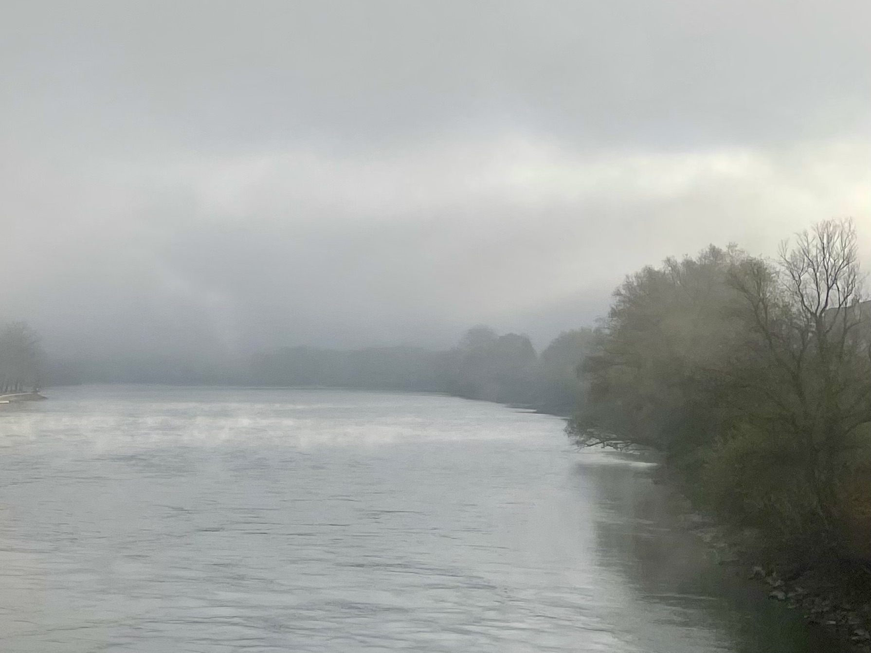 Das Foto zeit den Fluss Inn in Passau kurz vor dem Zusammenfluss mit der Donau. Am linken Ufer sind Bäume sichtbar. Der Morgennebel lichtet sich über dem Wasser