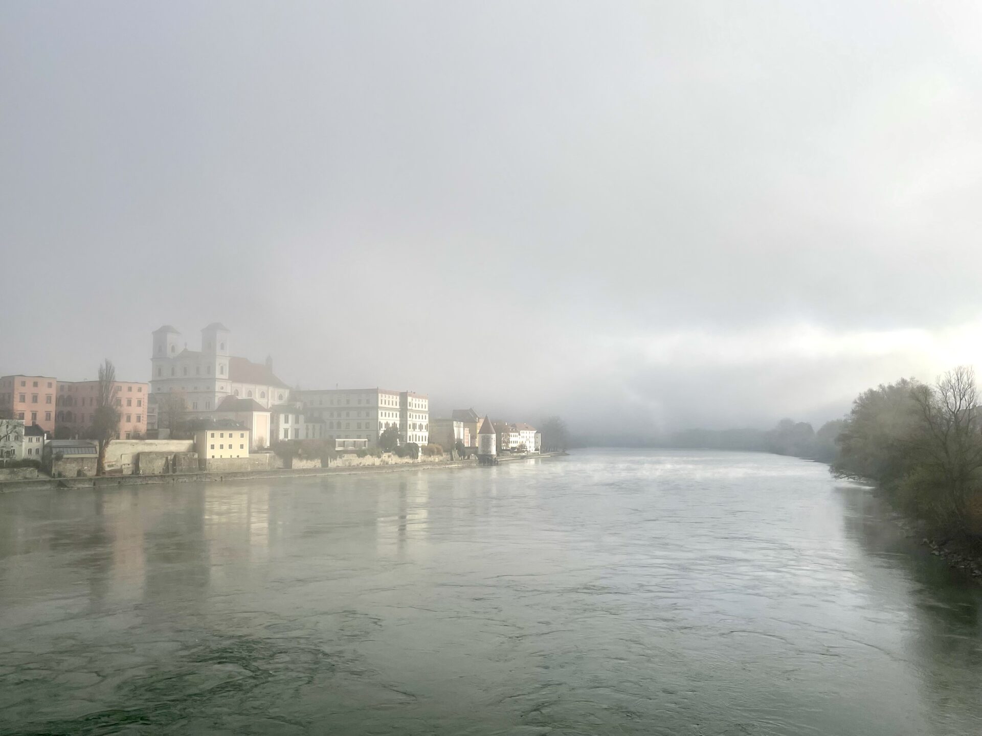 Das Foto zeigt den Inn in Passau. Auf der linken Uferseite sieht man Bäume, auf der rechten Uferseite liegt die Altstadt von Passau. Der Morgennebel wird von der Sonne aufgelöst.